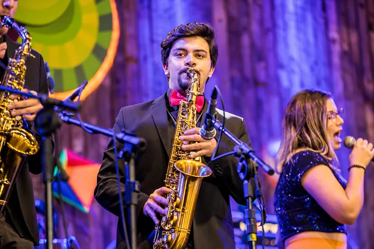 Victor Aguiar a tocar saxofone dourado numa festa privada animada, com cantora e brilhos coloridos ao fundo