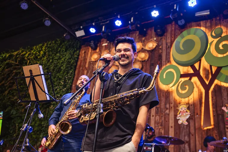 Victor Aguiar a cantar ao microfone com o saxofone, acompanhado por outros músicos num palco de madeira