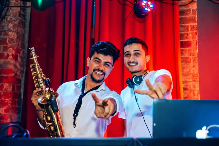 Victor Aguiar com o saxofone ao lado do DJ, num palco com cortinas vermelhas e luzes de festa
